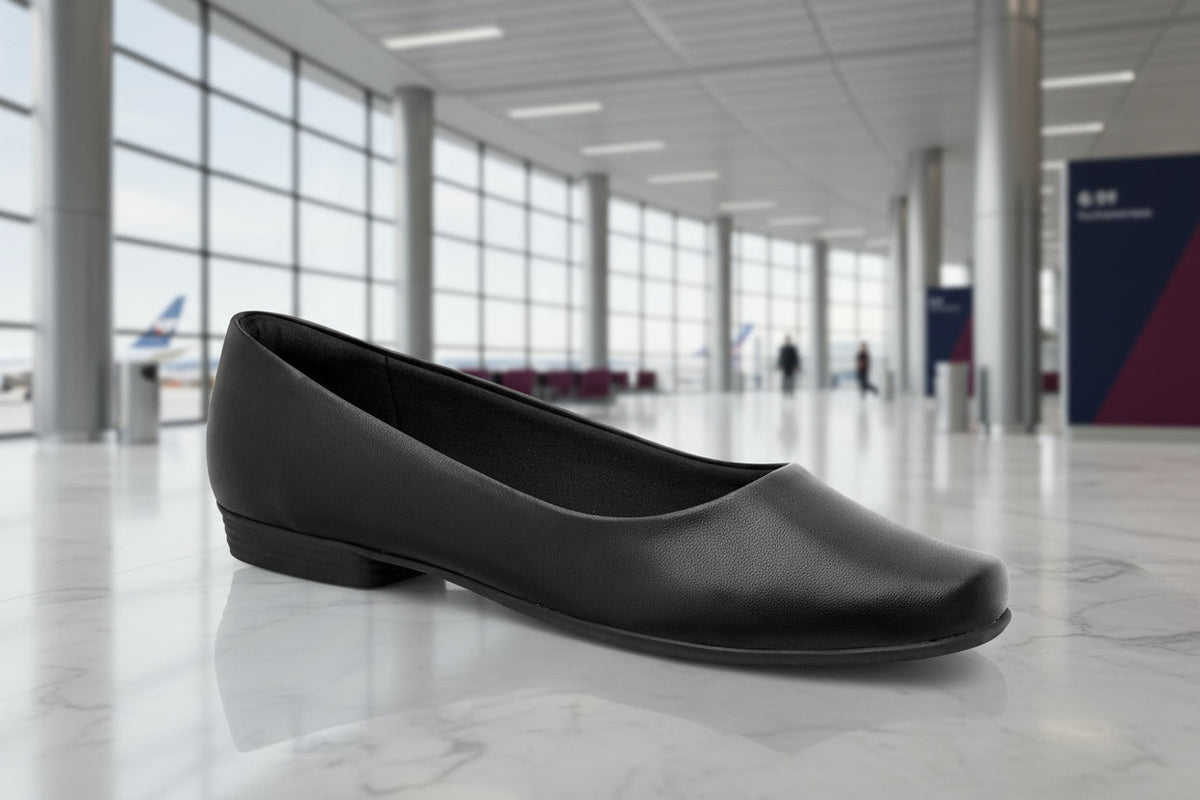 A single Piccadilly Ref: 250115-1052 Business Court Flat Shoe in Plane Black, a comfortable women's flat for flight attendants, rests on a shiny airport floor with large windows and blurred airplanes outside.